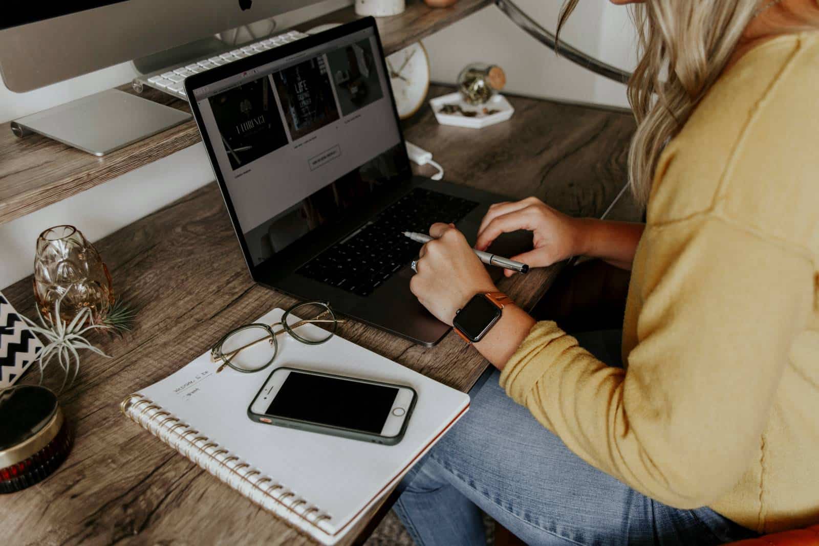 Woman sat at computer with notepad, pen and phone next to her.