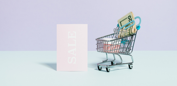 A trolley filled with cash, next to a sign that says SALE. This is on a pastel background