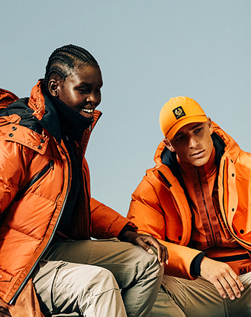 a man and woman wearing orange Belstaff jackets sat on rocks
