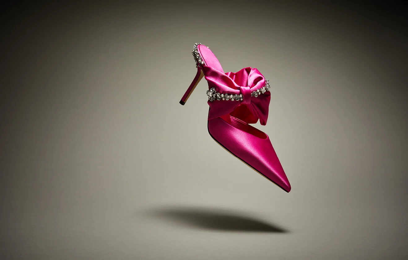 Pink satin Jimmy Choo mule with crystal embellishments floating in front of beige background.