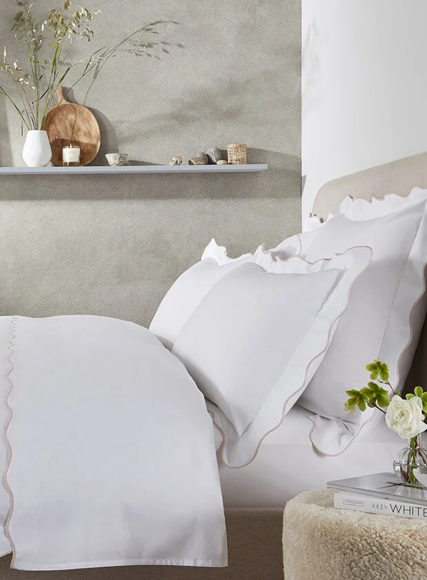 Side shot of a bed set up with white bedding featuring beige trim from The White Company.