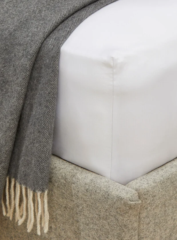 A close up of the corner of a white fitted bedsheet on a bed with a grey blanket draped to the side.