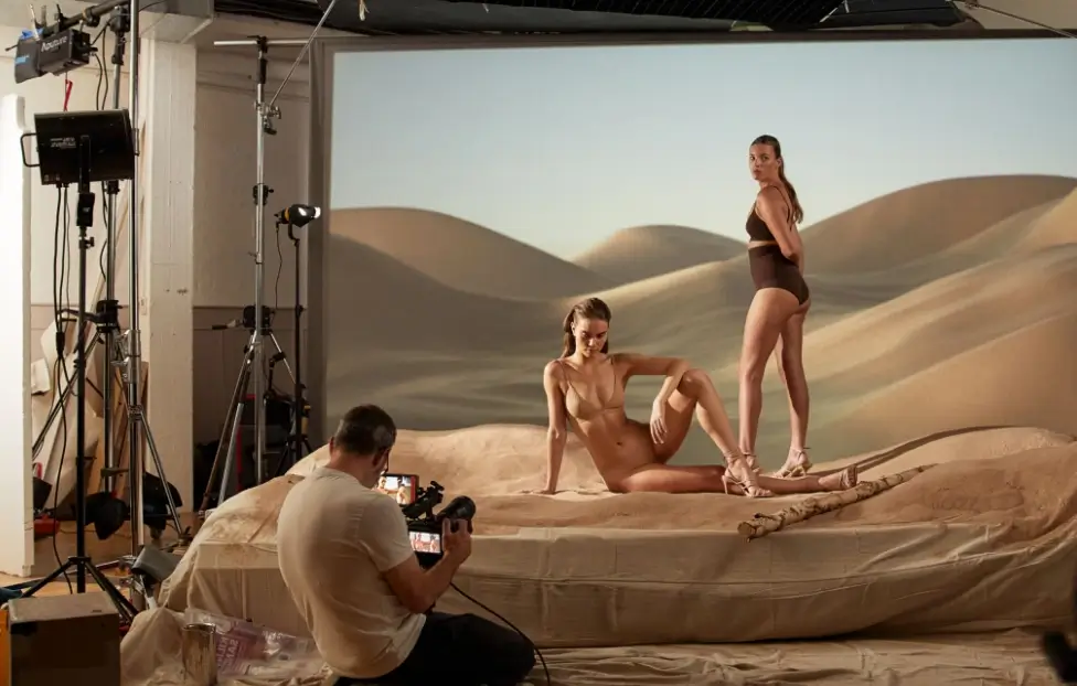 Two women posing on photography set in front of a desertscape image with photographer holding camera.