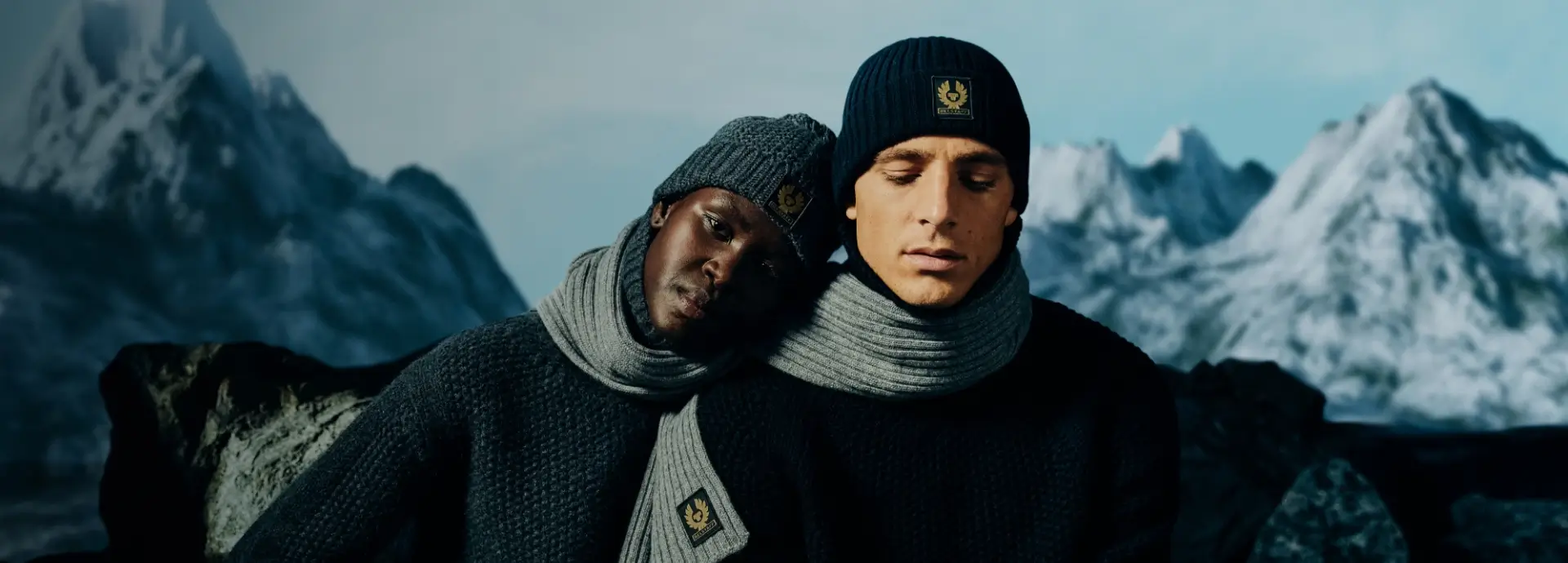Two models wearing hats and scarves leaning on each other in front of wintry landscape.