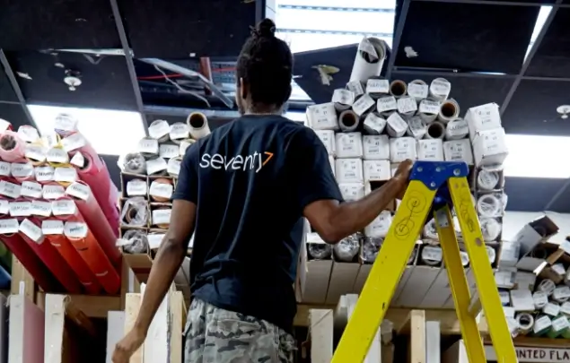 Member of Seventy7 staff up a step ladder looking at a stack of rolls of paper.