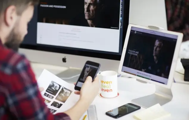 Person working on smartphone with laptop screen and desktop screen in front of them.