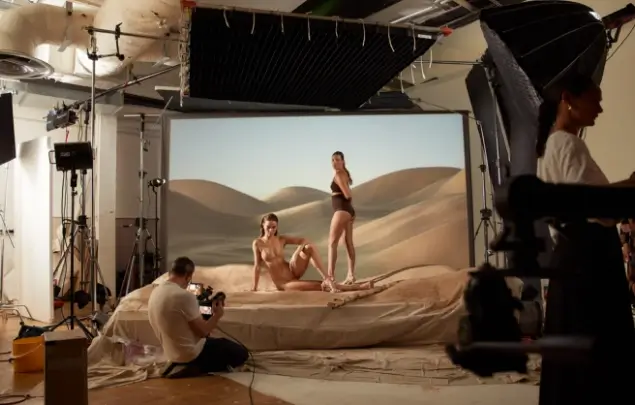 Two women posing on photography set in front of a desertscape image with photographer holding camera.