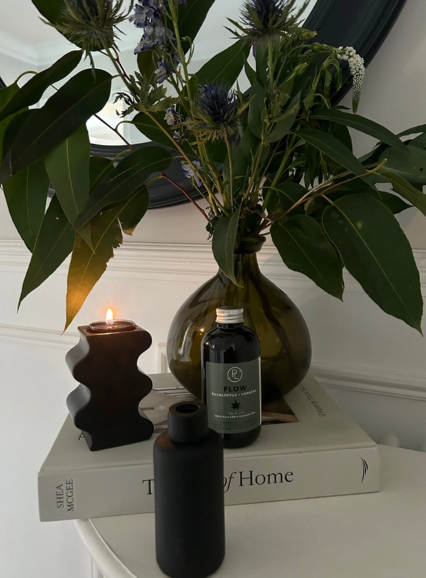 A monochrome display featuring PartyLite's refillable reed diffuser and tealight candle holder with a vase of green flowers and a book on home style.