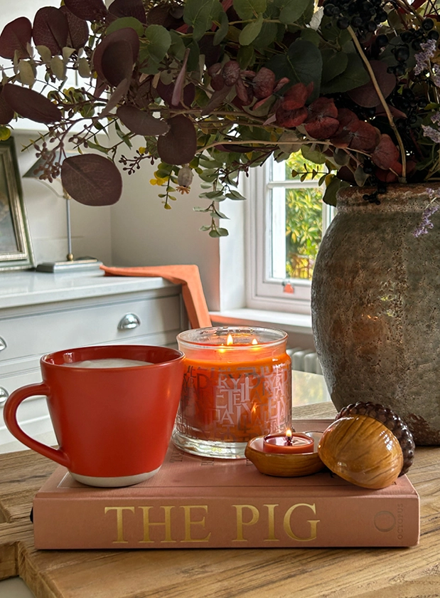 Large wooden table with vase full of greenery next to a hot drink in a mug and a lit PartyLite candle.